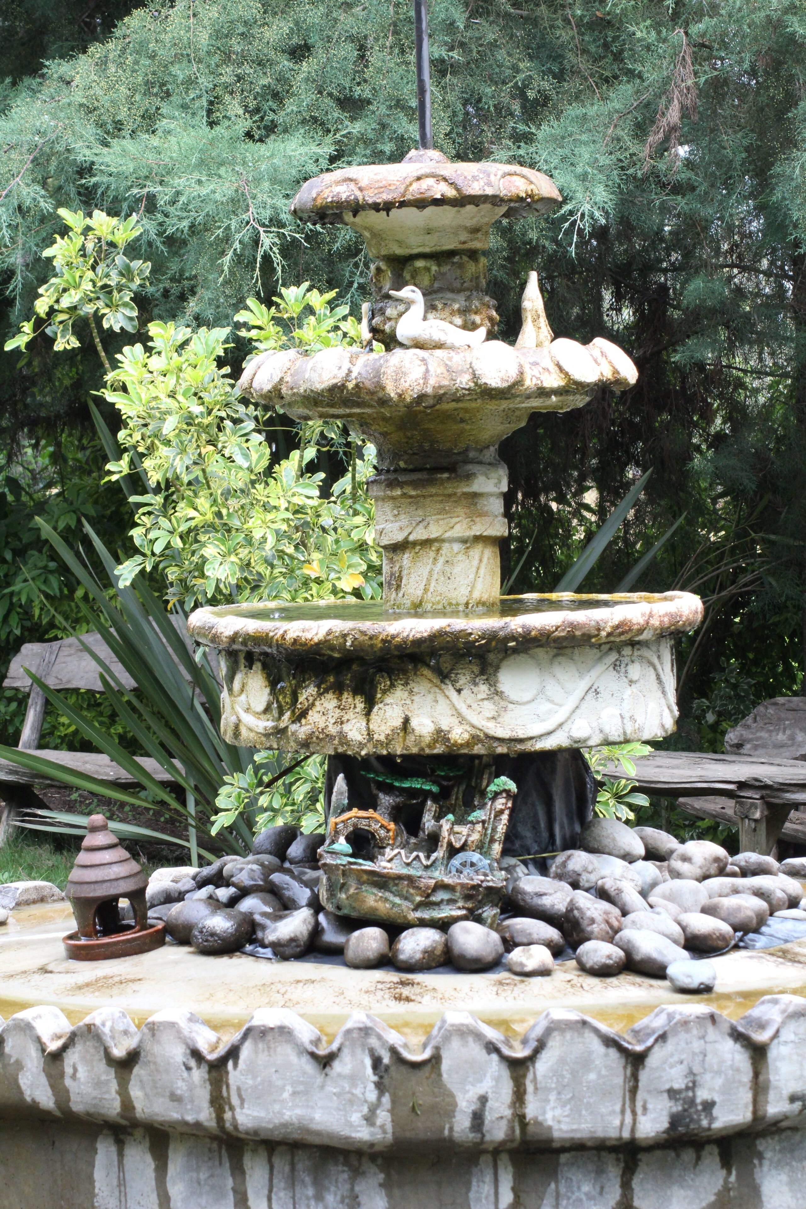 A multi-tiered stone fountain with a bird sculpture, surrounded by rocks and greenery.