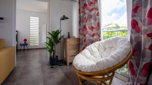 Un salon moderne avec un fauteuil en osier, une plante en pot, une commode en bois et une vue dégagée sur l’extérieur grâce à de grandes fenêtres.