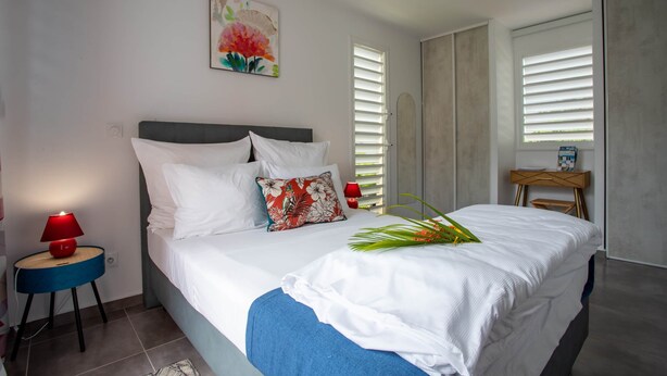 Un lit bien fait, avec des draps blancs et un plaid bleu, une lampe rouge posée sur une petite table de chevet en bois, et un tableau de fleurs accroché au mur.