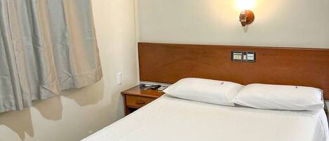 A hotel room with a bed, a nightstand, an air conditioning unit, a wall-mounted light, and a window with curtains.