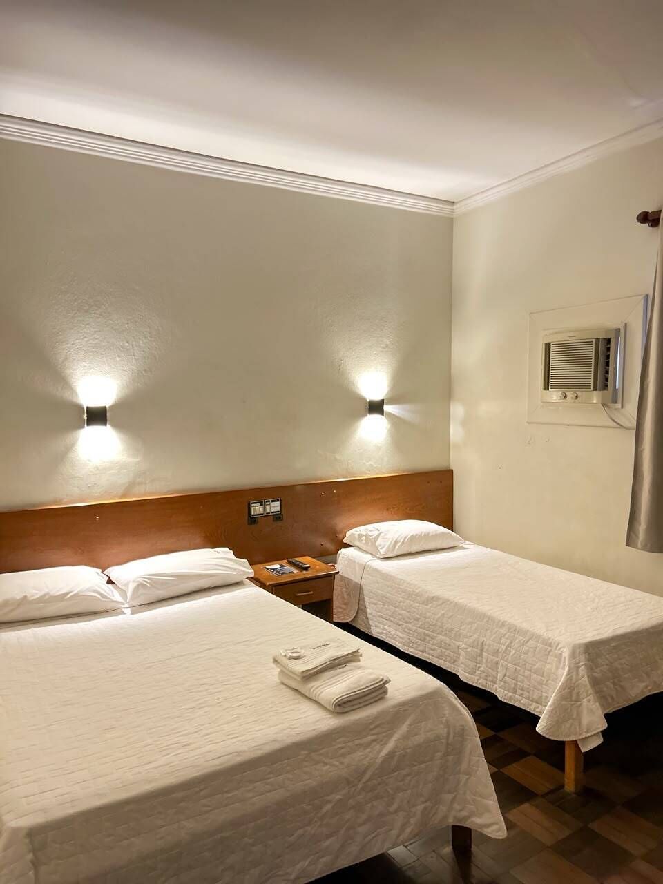 Habitación de hotel con dos camas individuales, un cabecero de madera, una mesita de noche, un calentador de pared y una ventana con persianas.