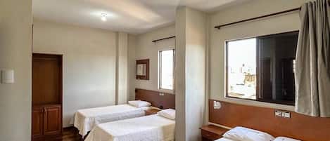 A hotel room with two single beds, a wooden wardrobe, a large window with curtains, and a ceiling light.