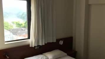 A hotel room with a bed, a nightstand, a window with curtains, and a view of greenery outside.