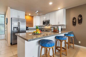 A modern kitchen with white cabinets, a stainless steel refrigerator, a microwave, and a granite countertop. There are two wooden stools with blue cushions and a ceiling fan. - KKSR#3103 DIRECT OCEANFRONT, INCREDIBLE OCEAN VIEWS & REMODELED! (Kailua Kona)
