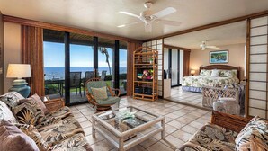 A spacious room with a large window overlooking the ocean, a bed with a floral bedspread, a wicker chair, and a glass coffee table.