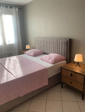 Ein ordentlich bezogenes Bett mit grauem Kopfteil, zwei Kissen und einer rosafarbenen Tagesdecke. Ein Nachttisch aus Holz mit Lampe und Schublade steht daneben.