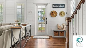 A room with a wooden floor, a sofa, a table with a vase and a plant, a door with a view to the outside, and a sign on the wall. - Cabana Banana - 4 Bikes / 6-Seater Golf Cart (Santa Rosa Beach)
