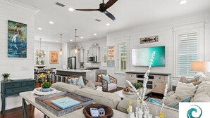 A spacious living room with a large TV, comfortable seating, and a kitchen in the background. - Cabana Banana - 4 Bikes / 6-Seater Golf Cart (Santa Rosa Beach)