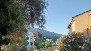 A narrow cobblestone street with a parked motorcycle and a car on the left. There are buildings on both sides, and a hill with a church in the background. - Studio Climatisé Possibilité 4 Adultes/parking Privé (grasse)