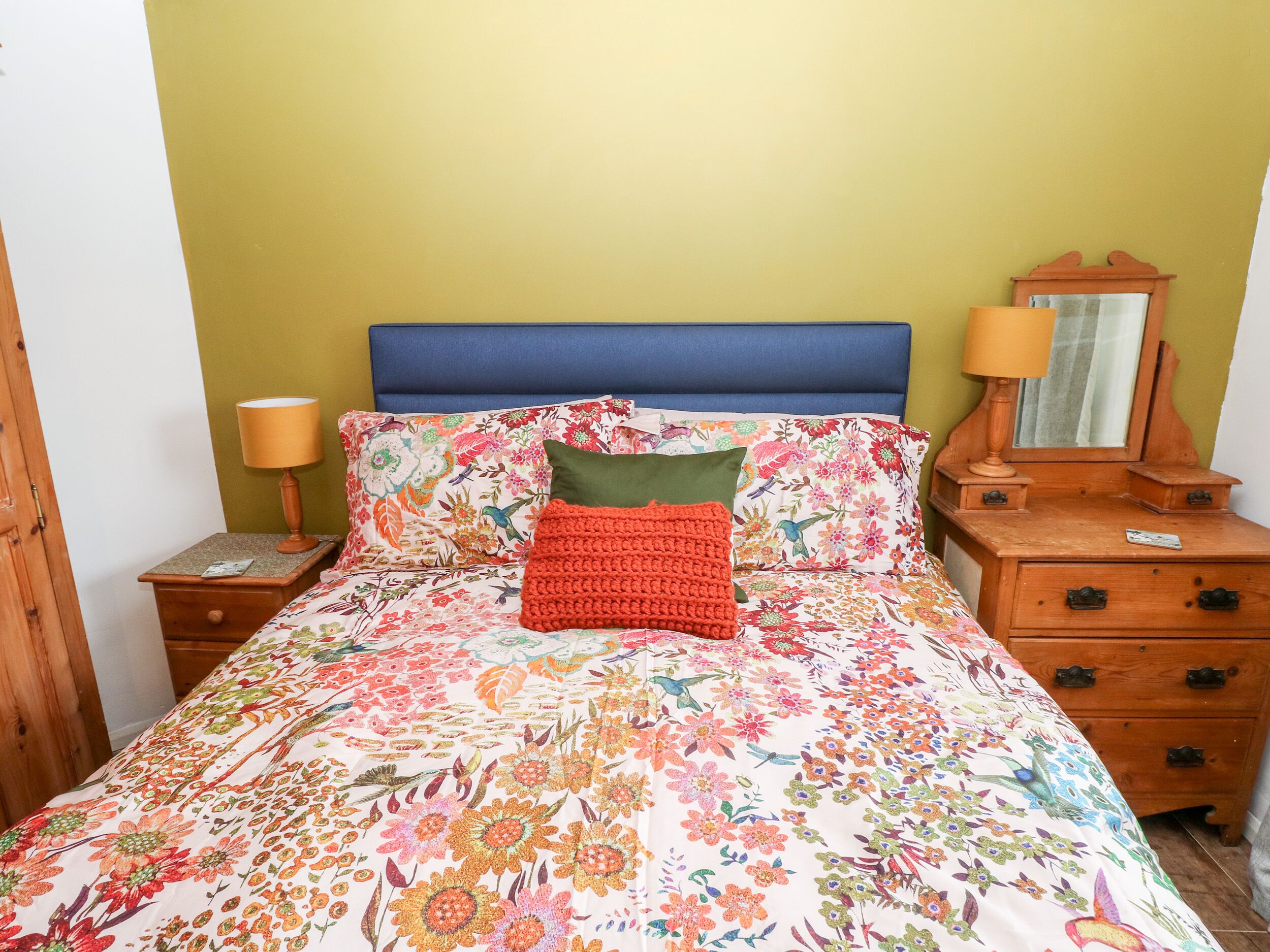 A bed with floral bedding, a blue headboard, and a wooden nightstand with a lamp.