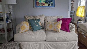 Un salon avec un canapé beige, des coussins colorés, un tableau encadré et une bibliothèque.