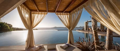 Un área de descanso al aire libre iluminada por el sol, con una terraza de madera, dos zonas de estar con cojines y vistas a una gran masa de agua.