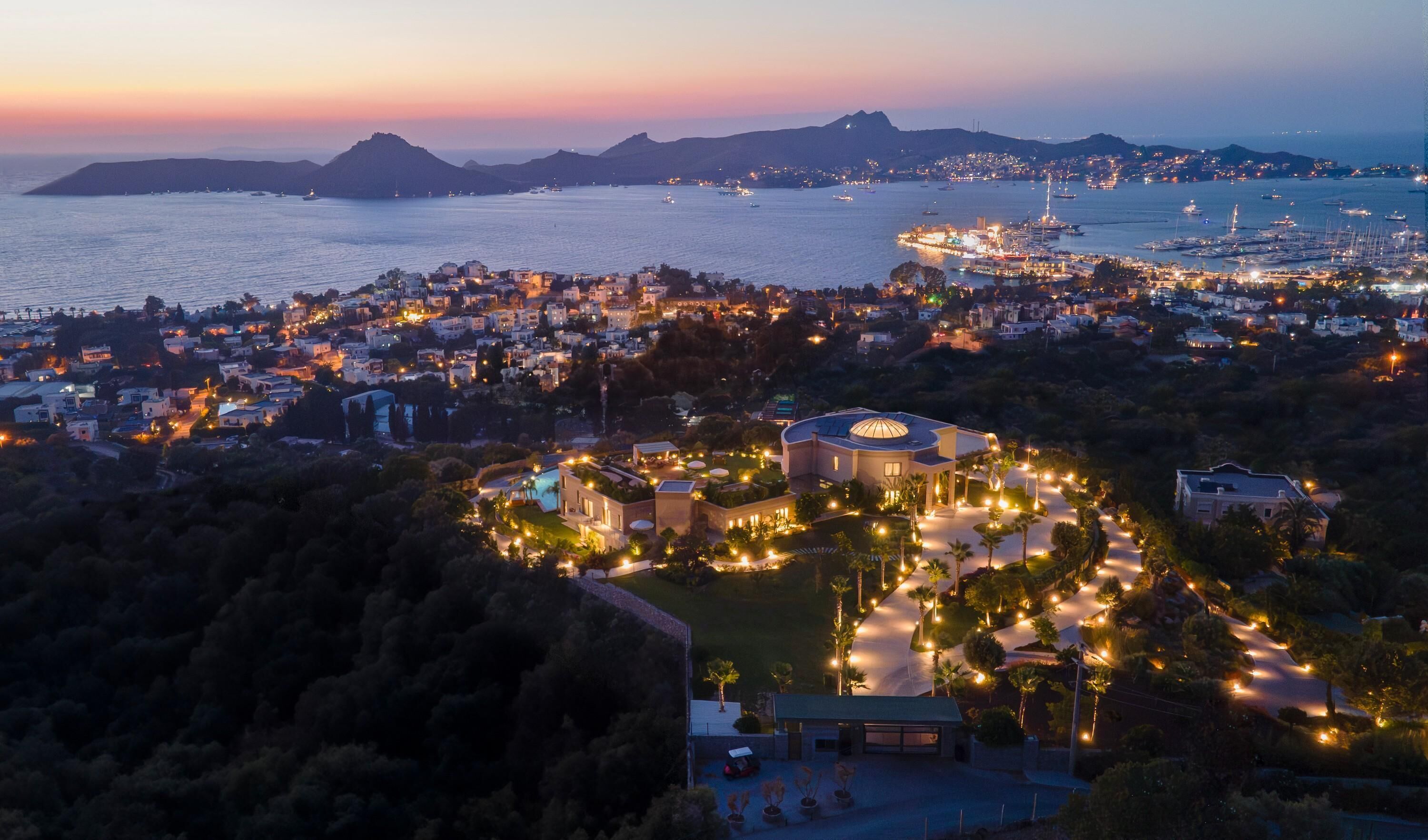 Une ville côtière au crépuscule, avec un imposant bâtiment circulaire entouré de rues bien éclairées et de verdure.