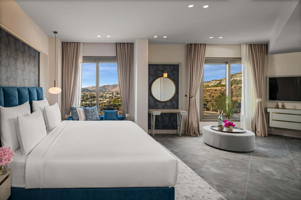 A modern hotel room with a large bed, a round mirror, a TV, and a view of the city.
