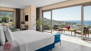 Habitación de hotel moderna con una cama grande, un balcón con vistas a la ciudad y al mar, un televisor de pantalla plana y una pequeña mesa con una planta.