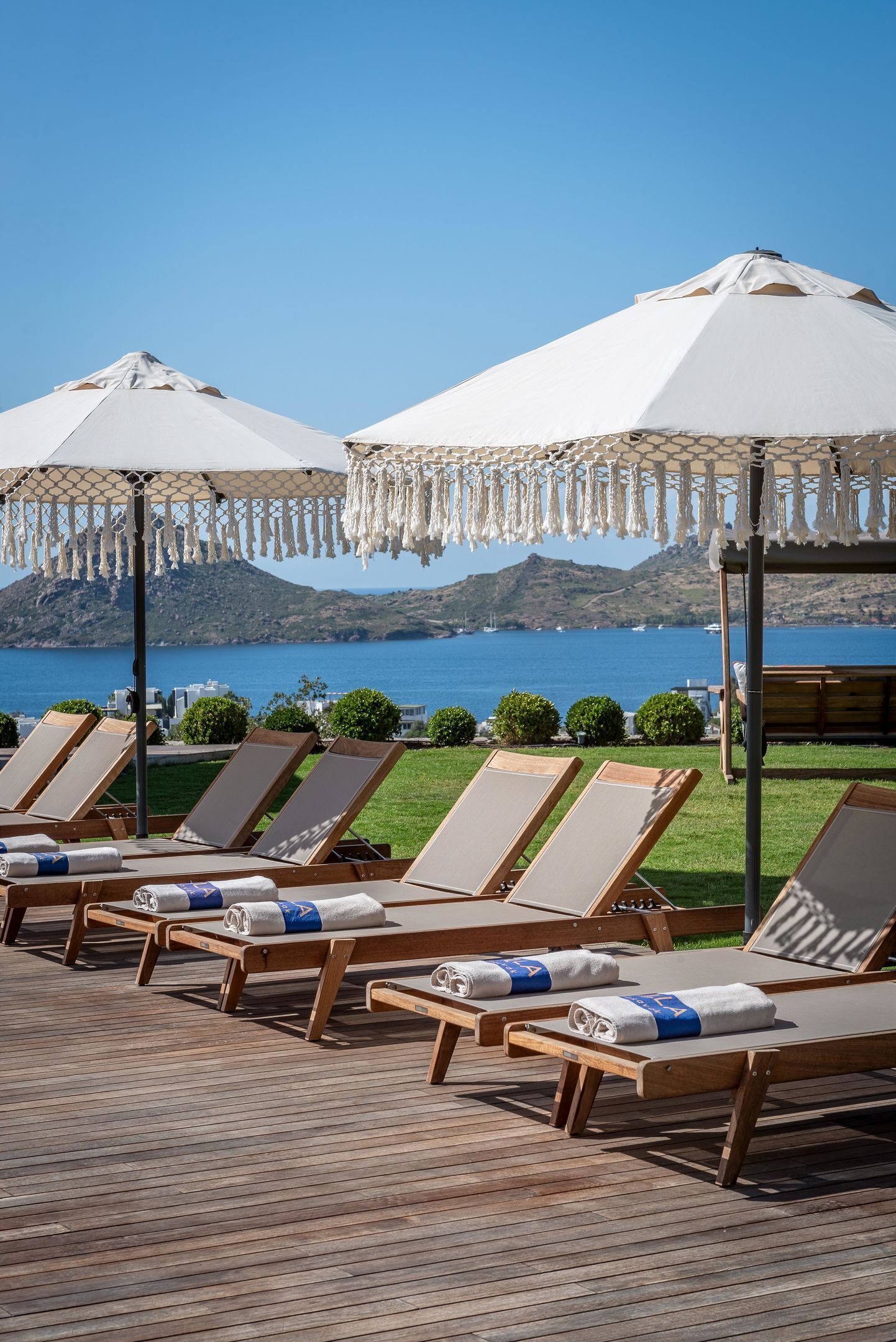 Une rangée de chaises longues en bois, avec des serviettes, des parasols blancs, et une vue sur un lac et des collines.