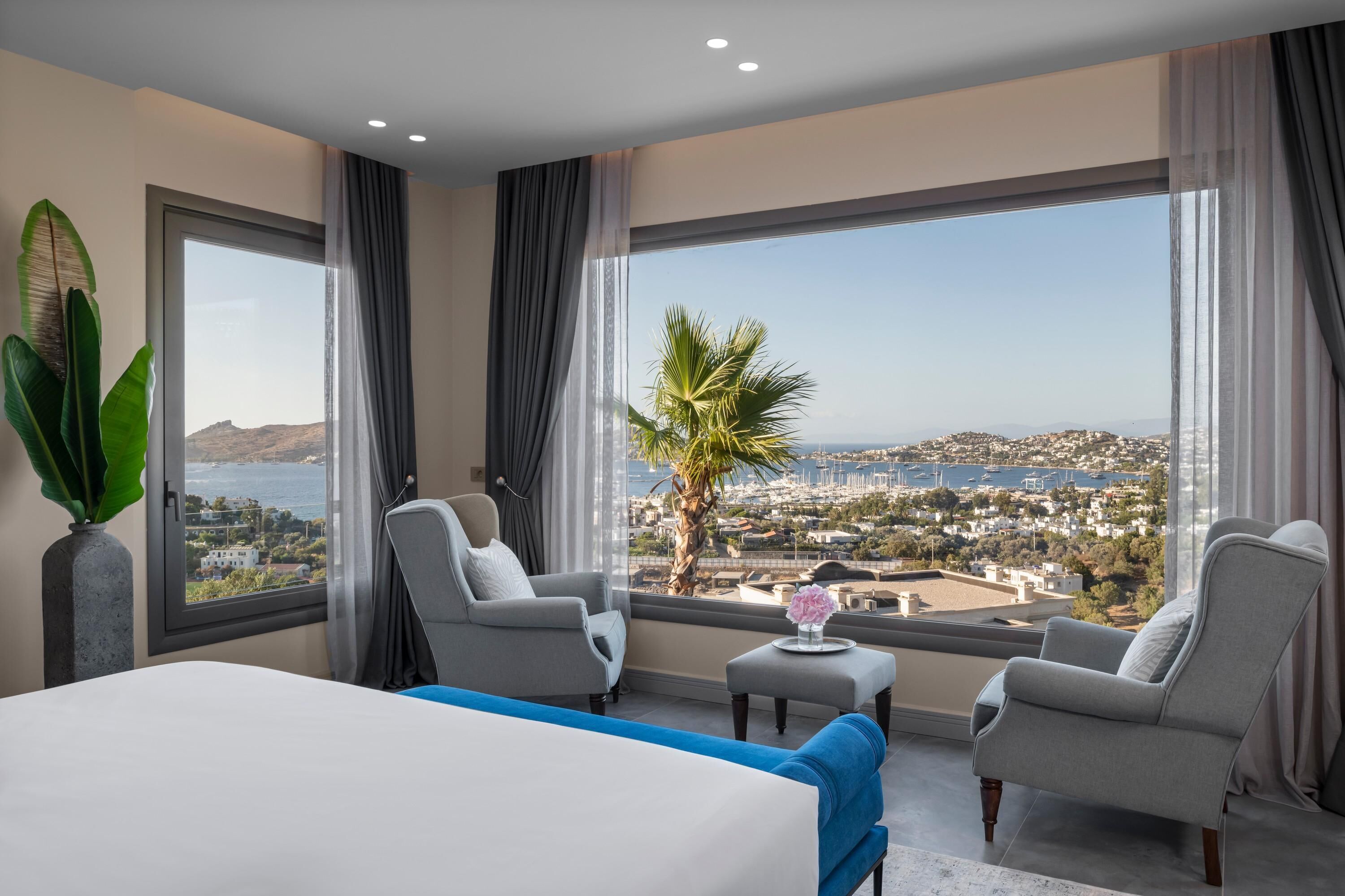 Una habitación de hotel con una cama grande, dos sillones, una mesita con un jarrón y vistas al mar y a la ciudad.