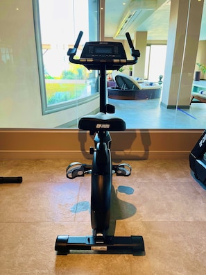 An exercise bike with a digital display, positioned indoors with a view of an outdoor patio area through a large window. - Ailla Yalıkavak (Bodrum)