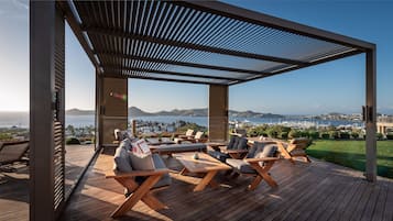 Un espacio de estar al aire libre cubierto con mobiliario de madera y vistas al mar y al paisaje urbano.