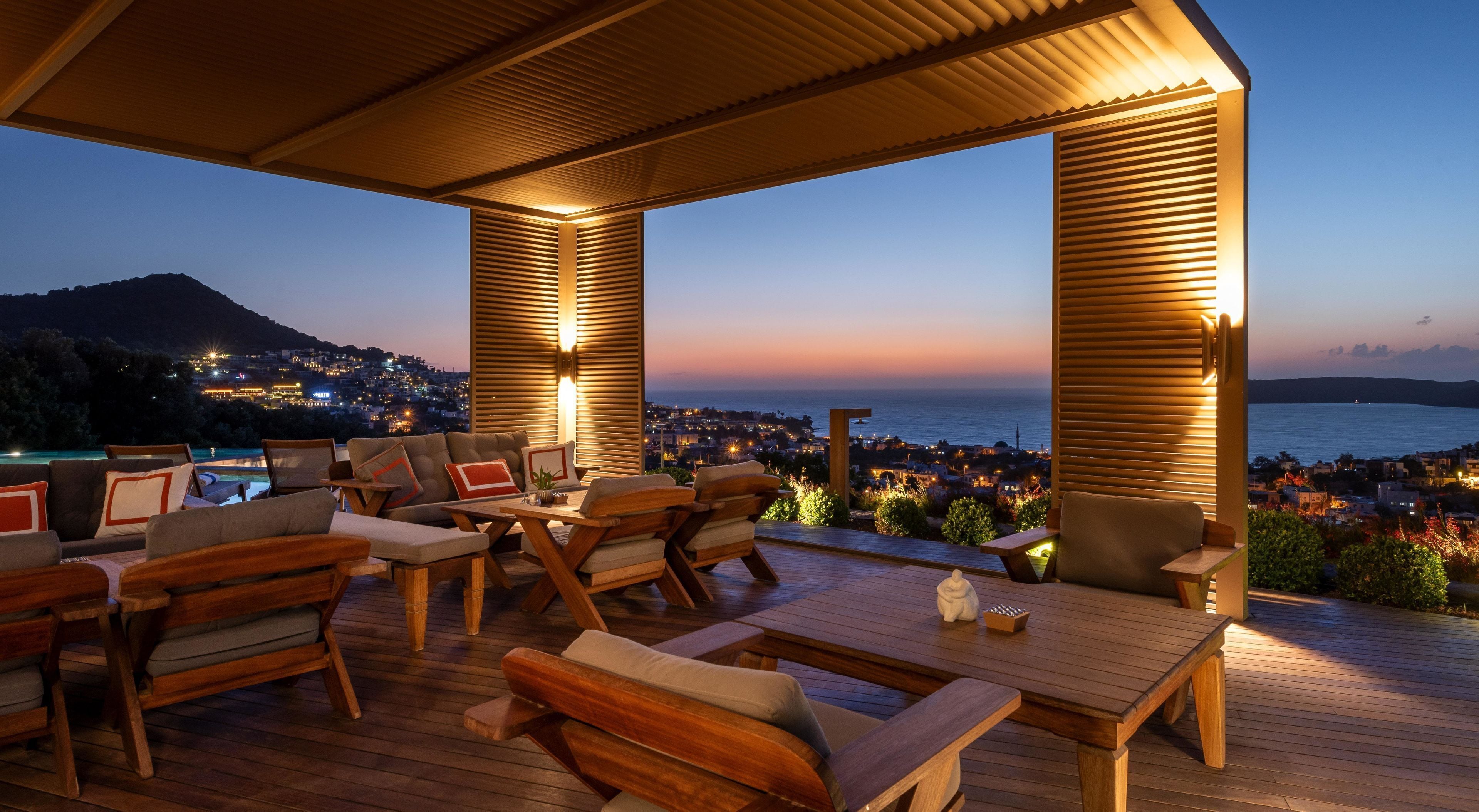 Una terraza con mobiliario de madera, piscina y vista a una ciudad al atardecer.
