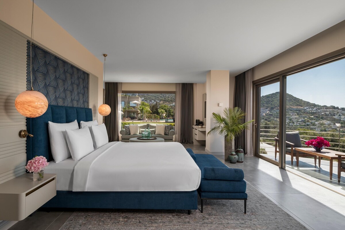 A modern bedroom with a large bed, a view of the city, and a balcony with seating.