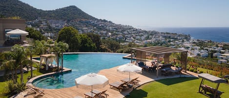A pool area with lounge chairs and umbrellas, surrounded by a wooden deck and a grassy lawn, overlooking a coastal town and hills.