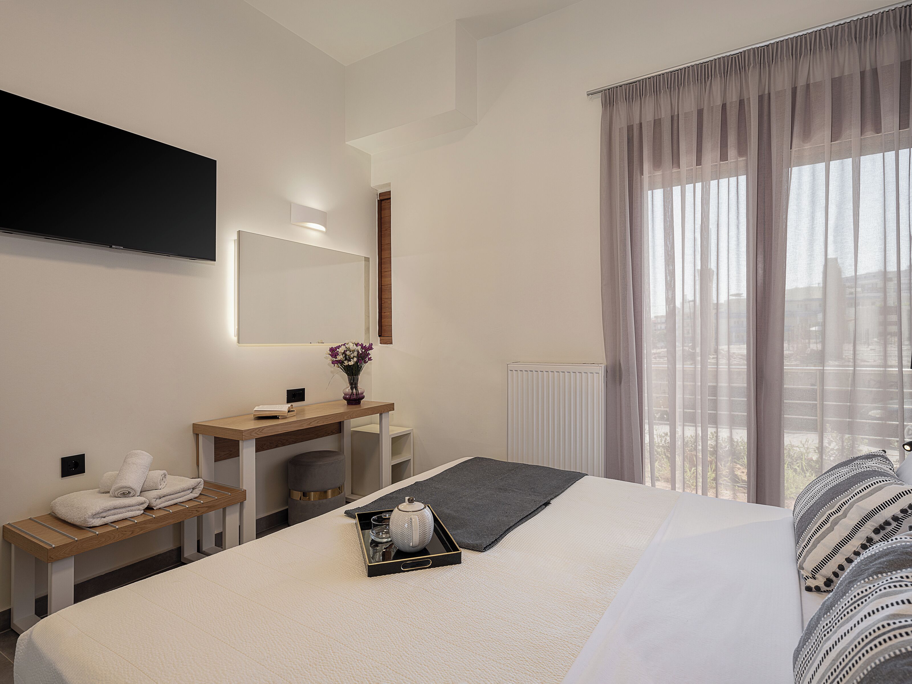 A modern hotel room with a large bed, a bedside table with a vase of flowers, a flat-screen TV mounted on the wall, and a window with sheer curtains.