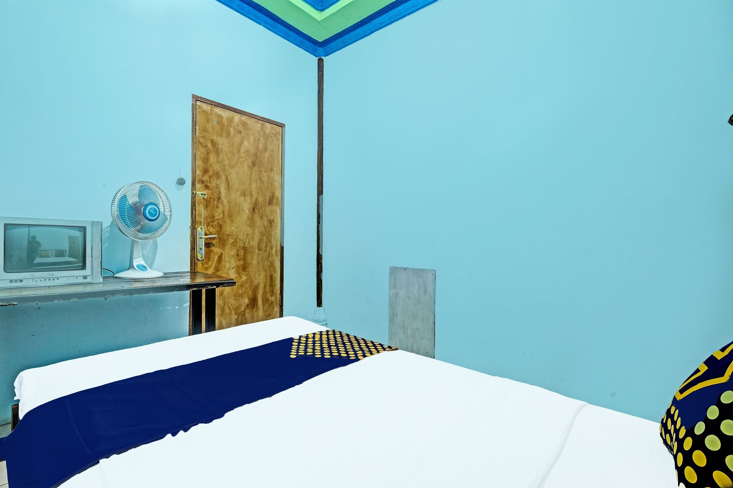 A room with a bed, a wooden door, a desk with a computer and a fan, and a blue wall.