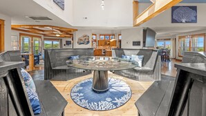 Un espacio habitable amplio con una mesa de vidrio central rodeada de sofás oscuros, vigas de madera en el techo y una vista de la cocina al fondo.