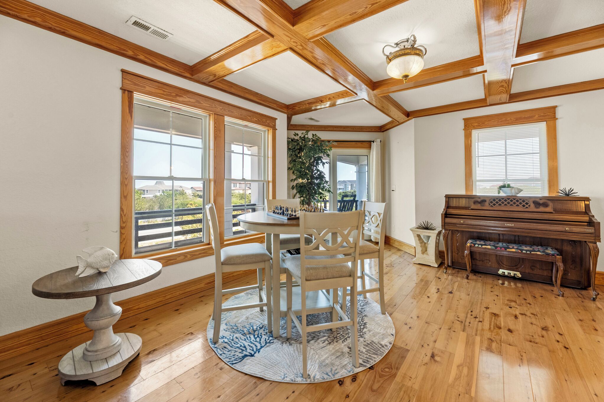 A dining area with a round table, chairs, a piano, and a view through large windows.