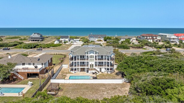 Una zona residencial costera con una casa grande de varios pisos que cuenta con una piscina, rodeada de casas más pequeñas y una vista despejada del océano.