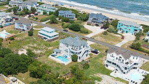 Oversigtsbillede af et kystnære boligområde med flere huse, et stort hus i midten med swimmingpool, og en strand i baggrunden.