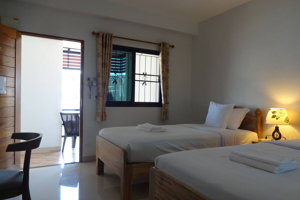 Une chambre d’hôtel avec deux lits, une chaise en bois, une table, une fenêtre avec des rideaux et une lampe.