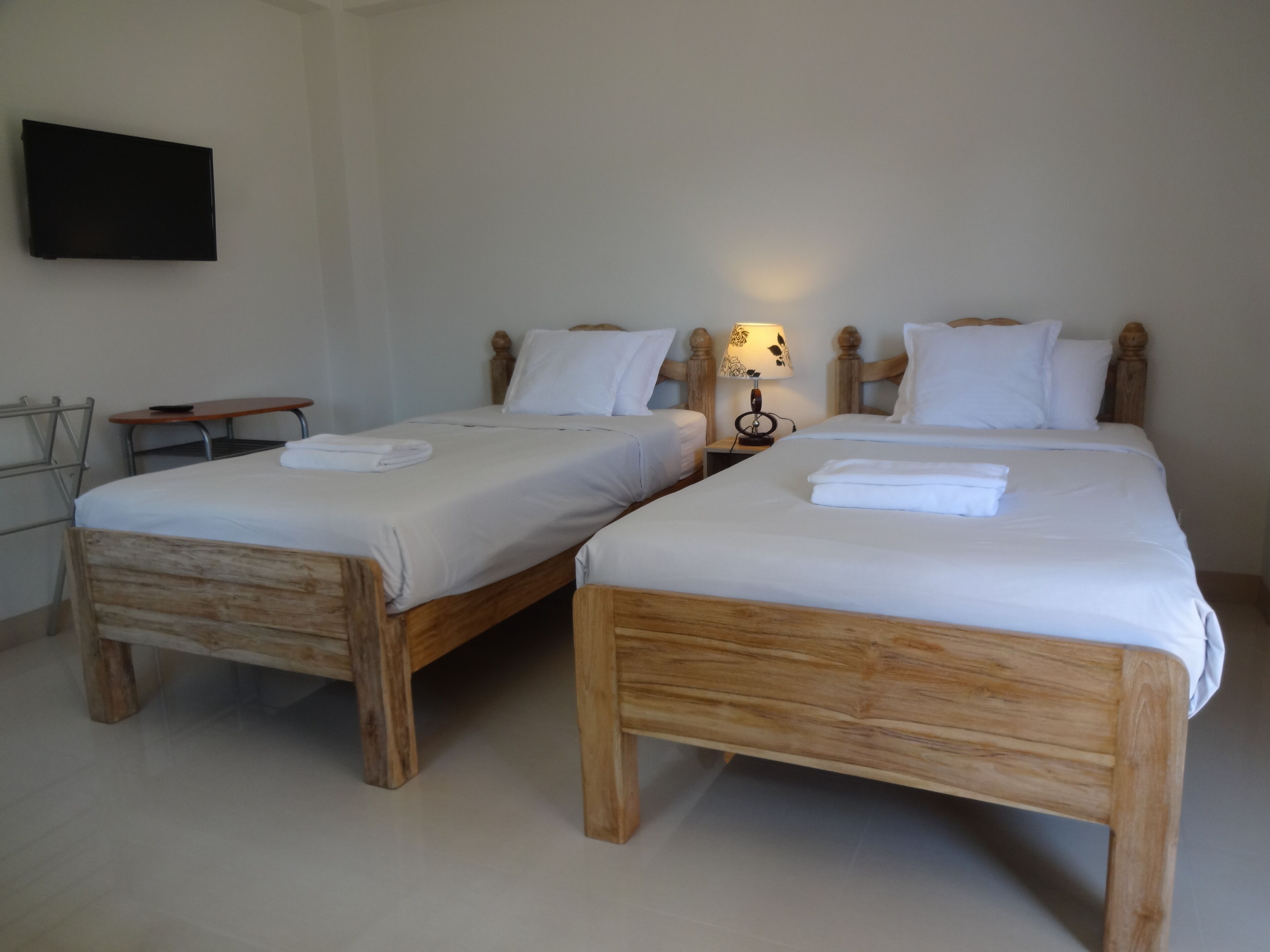 Two wooden beds with white linens, a small bedside table with a lamp, and a flat-screen TV mounted on the wall.