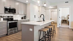 A modern kitchen with stainless steel appliances, a white island with stools, and pendant lighting. - Lagoon Breeze: Desert Color Lagoon + Beaches (St. George)