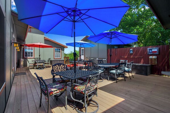 An outdoor patio with multiple tables and chairs, blue umbrellas, and a wooden deck. - Swing On Inn I Hot tub, Sleeps 12 (Dripping Springs)