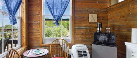 Una habitación con paredes de madera, una ventana con cortinas azules, una silla, una mesa con revistas y un refrigerador.