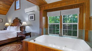 A bedroom with a large bed, a wooden headboard, a window with a view of trees, and a white bathtub.