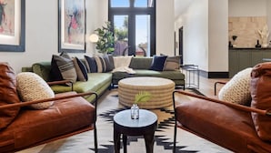 A modern living room with a sectional sofa, two leather armchairs, a round ottoman, and a coffee table. There is a large window with a view of the outdoors, framed artwork on the walls, and a kitchen area in the background. - Stunning, Designer Historic Home | Heated Pool (New Orleans)
