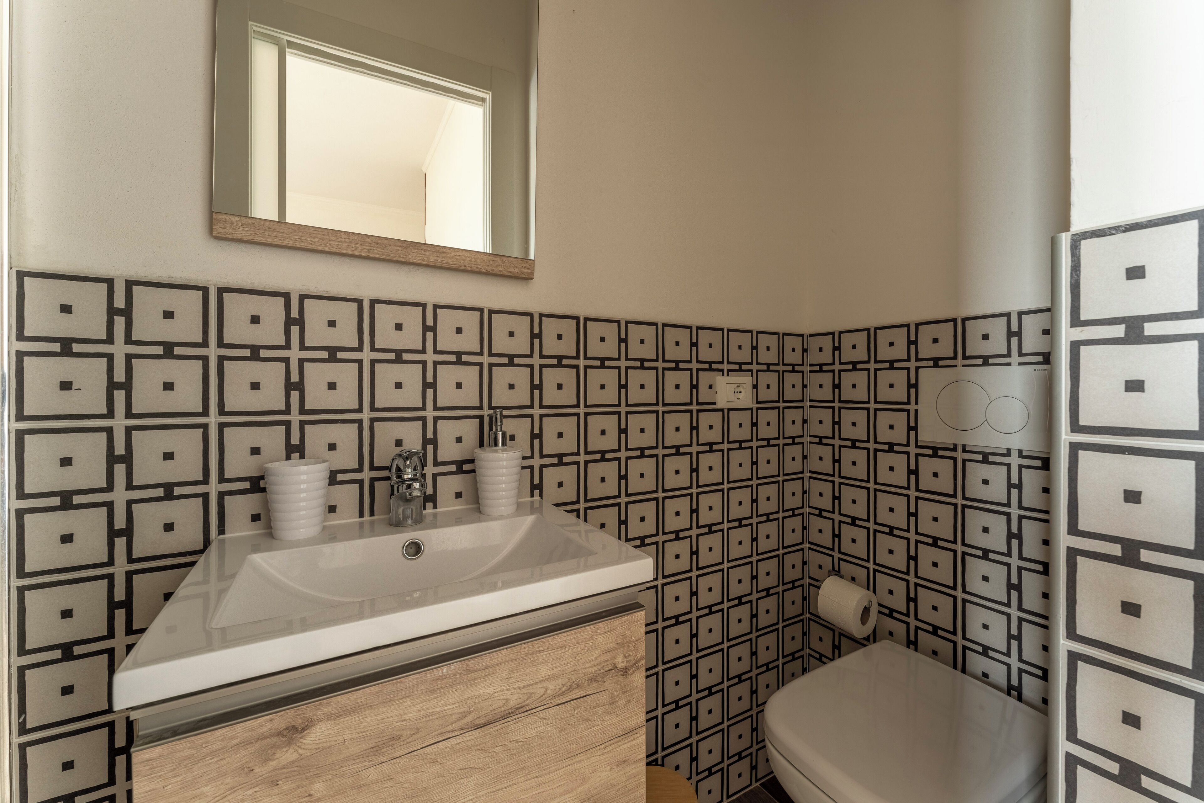 A bathroom with a sink, mirror, and toilet. The walls are tiled with a geometric pattern.