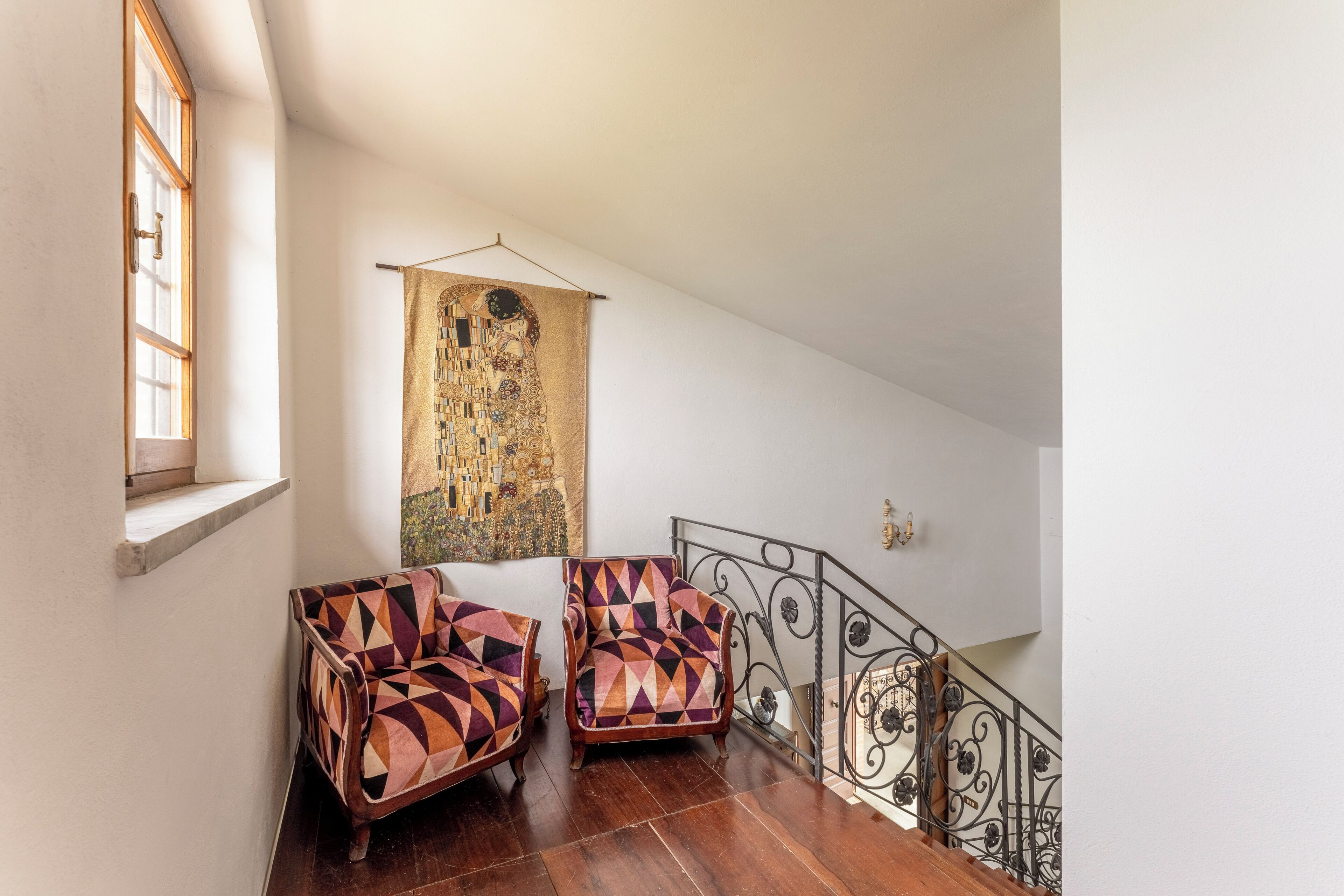 A staircase with a wrought iron railing, two patterned armchairs, and a large tapestry on the wall.
