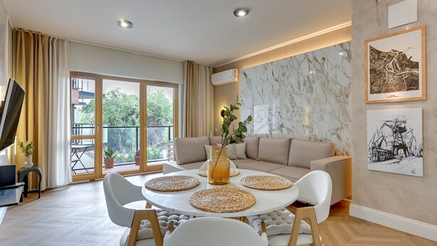 A modern living room with a round table, white chairs, a beige sofa, and artwork on the walls. - RentPlanet - Sienna Grobla (Gdansk)