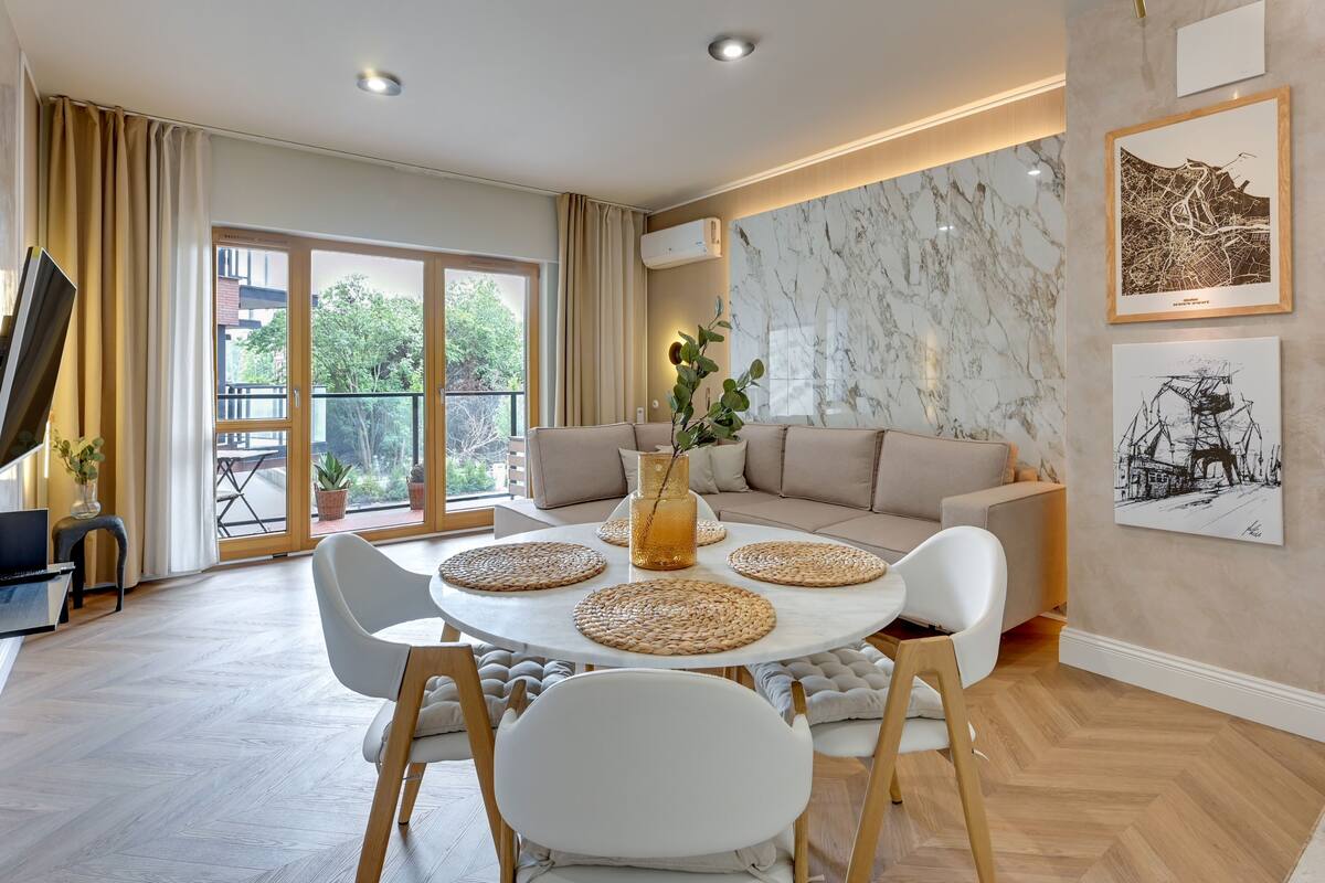 A modern living room with a round table, white chairs, a beige sofa, and artwork on the walls.