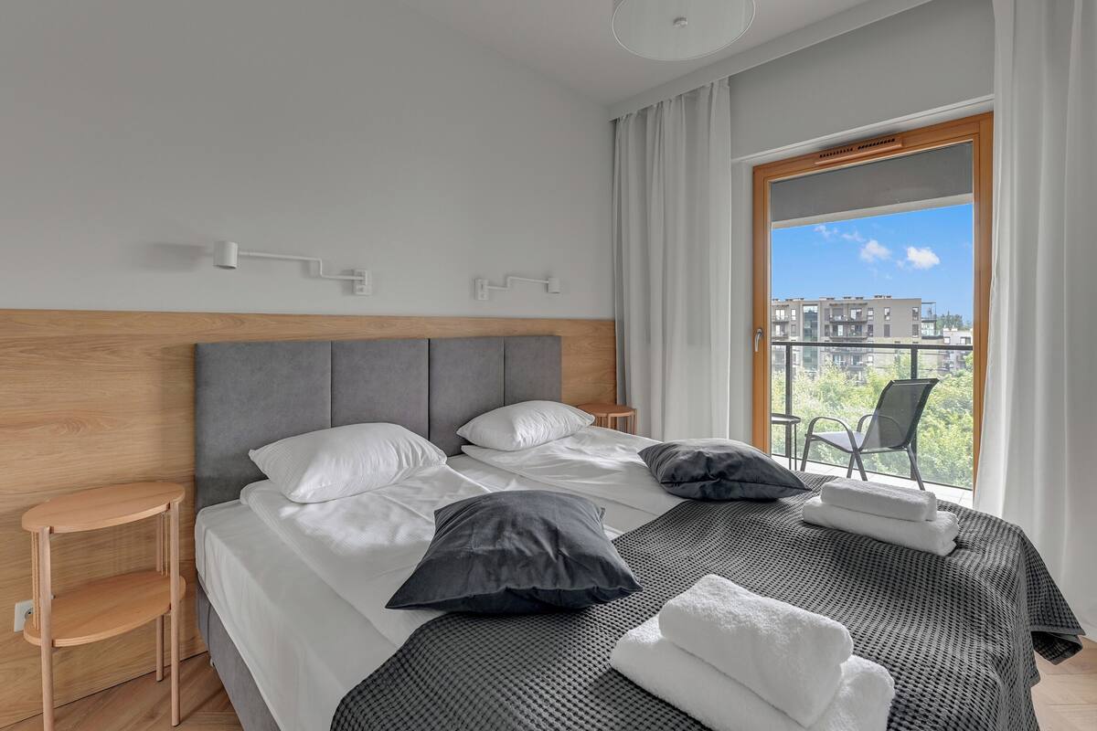 A neatly made bed with white and gray bedding, a wooden headboard, and a view of buildings through a large window.