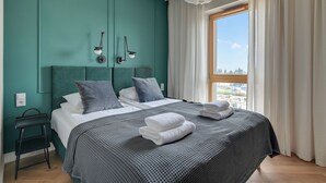 A modern bedroom with teal walls, a large bed with grey bedding, two bedside tables, and a view of the city through a window with white curtains. - RentPlanet - Sienna Grobla (Gdansk)