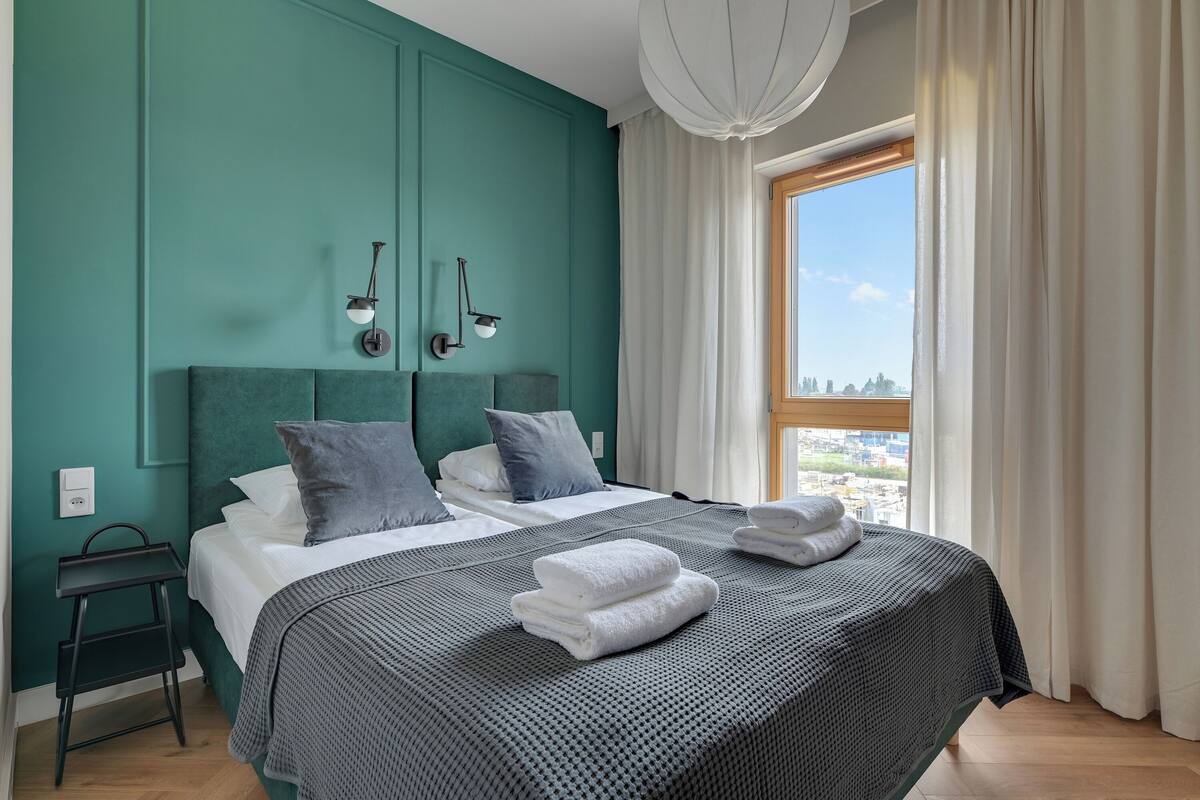 A modern bedroom with teal walls, a large bed with grey bedding, two bedside tables, and a view of the city through a window with white curtains.