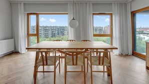A modern dining area with a wooden table and chairs, large windows with curtains, and a view of buildings and greenery outside. - RentPlanet - Sienna Grobla (Gdansk)