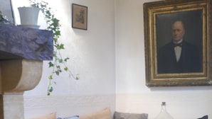 Un salon avec un canapé, des coussins, un pouf en osier, un portrait encadré accroché au mur et une plante en pot.