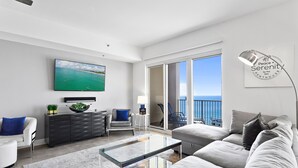 Ein modernes Wohnzimmer mit einem großen Fernseher, einem Glastisch und einer Couch. Ein Balkon bietet Blick auf das Meer.