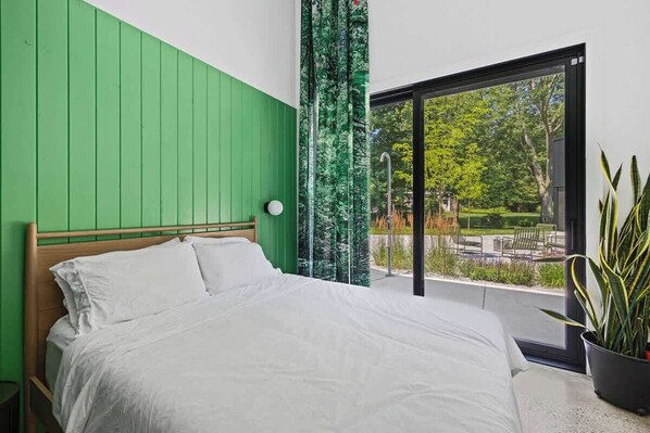 A bedroom with a green wall, a large bed, a window with a view of greenery, and a potted plant.